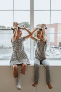 Zwei Kinder auf einer Fensterbank, schauen sich lachend an und halten mit ihren Händen Bücher über ihren Köpfen. 