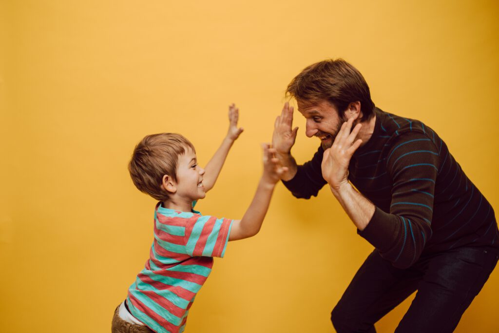 Ein Vater und sein Sohn stehen sich lachend vor einer gelben Wand gegenüber und geben sich ein High Five. 