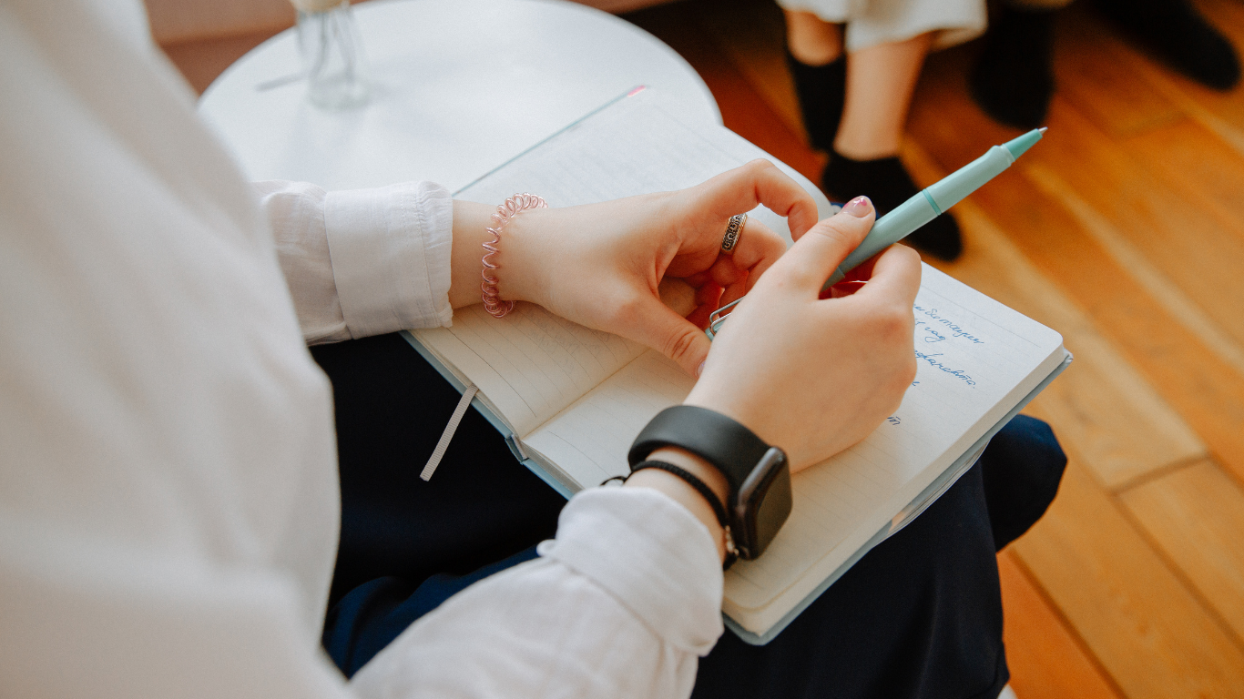 Eine Person einer Familienberatung schreibt Notizen in ein aufgeschlagenes Notizbuch, welches auf ihren Knien liegt. Sie hält einen Stift in der Hand.