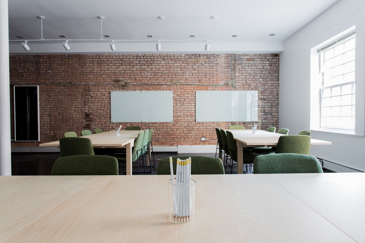 Ein Seminarraum mit Backsteinwand, davor zwei Tischen mit grünen Stühlen. Vorne im Bild stehen Stifte in einem Glas. Das Wort "Fortbildungen" ist zu sehen.