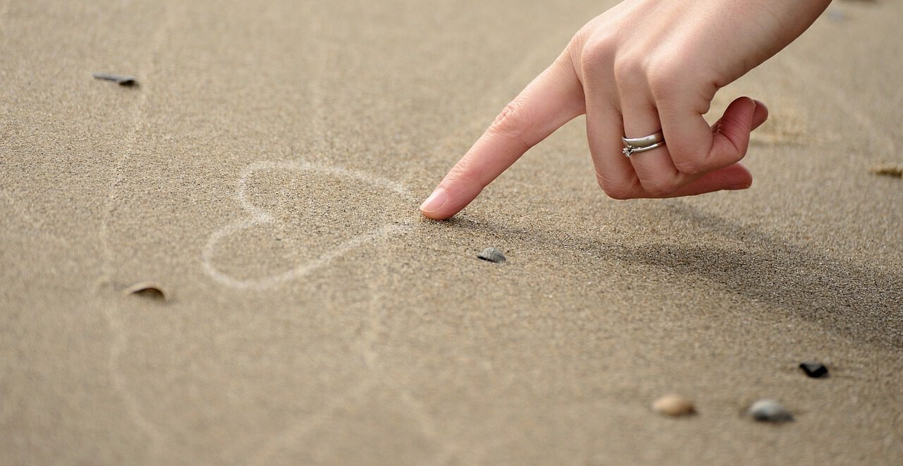 Eine Hand über Sand, die mit einem Finger ein Herz in den Sand gemalt hat. 