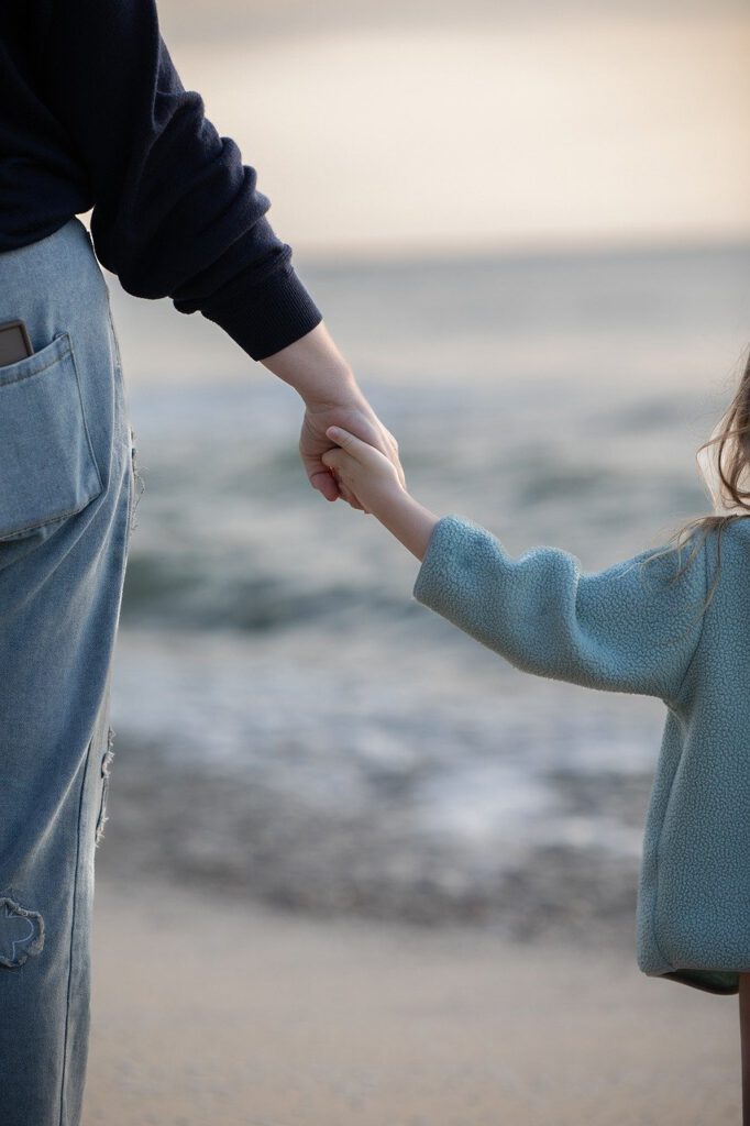 Mutter und Kind Hand in Hand am Strand, nur die Arme sind von hinten zu sehen. Symbolbild für Familie und Familienberatung.